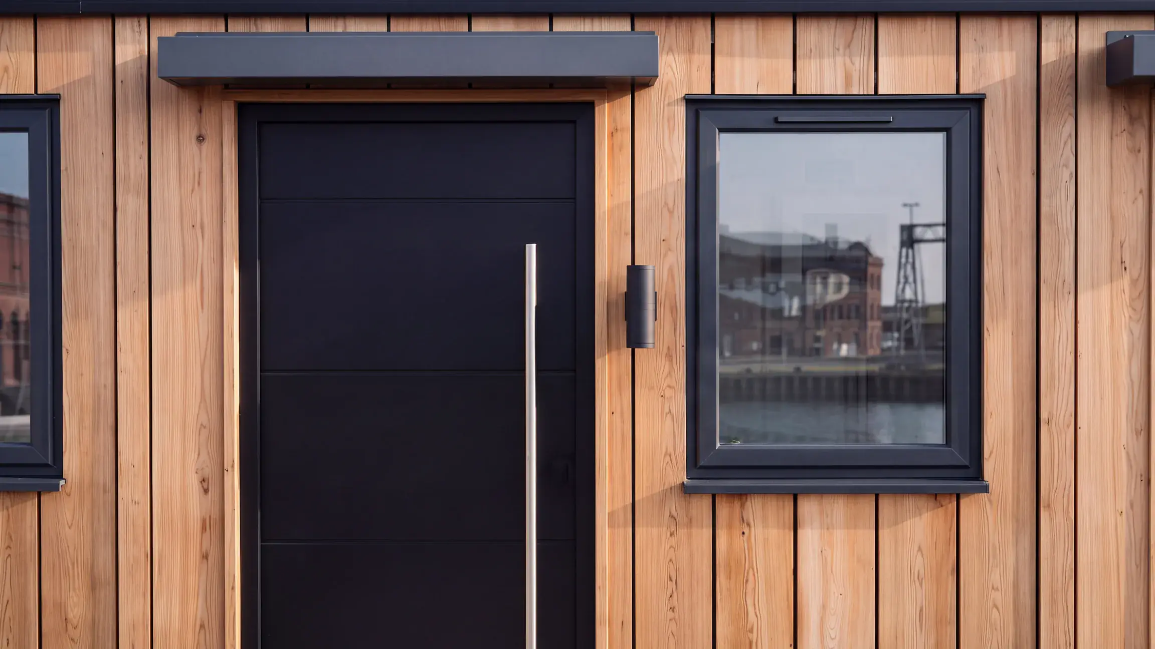 Close-up of a meanwhile home's front: a dark front door with a long steel handle under a small canopy, a wall light, and a window reflecting old brick buildings, set in vertical timber cladding.