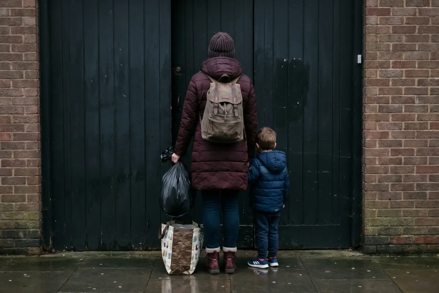Seen from behind, a mother with a backpack and bags of belongings stands with her young son in front of a shut black door.