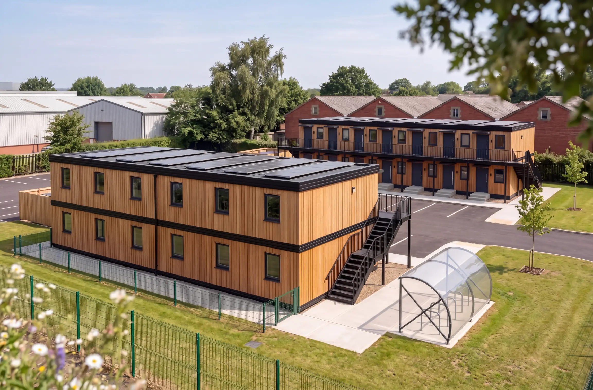 Two blocks of timber-clad Meanwhile homes with solar panels, one with an access deck and stair, beside a cycle shelter, new parking bays and young trees, with brick buildings, an industrial shed and mature trees behind.